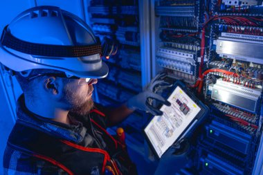 Elektrikçi, sigortalı santralde tablet kullanarak çalışmaya odaklanmış. Arka planda acil durum ışıkları olan karanlık odada kask takan özel kıyafetli elektrik mühendisi. Üst görünüm.