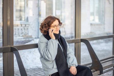 Otobüs durağında otobüs bekleyen ve telefonda konuşan bir genç. Toplu taşıma. Yüksek kalite fotoğraf. Toplu taşıma bekleniyor. Soğuk hava.