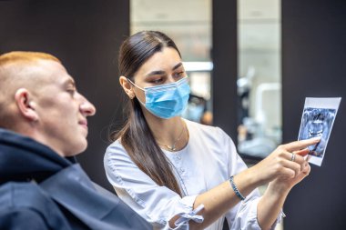 Genç bir dişçi hastaya diş röntgenini gösteriyor. Dişçi konsepti. Yüksek kalite fotoğraf. İşte bir dişçi. Dişçi ve hasta. Bir kadının yüzünde maske.