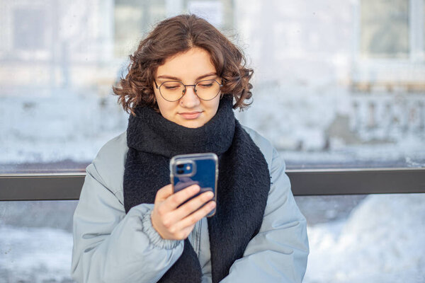 Teenage girl waiting for public transportation. Girl at the bus stop. High quality photo. Cold weather. A student girl is waiting for the bus. Phone in hand.