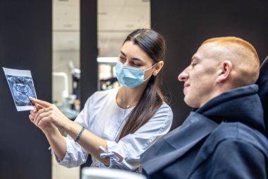 Genç bir dişçi hastaya diş röntgenini gösteriyor. Dişçi konsepti. Yüksek kalite fotoğraf. İşte bir dişçi. Dişçi ve hasta. Bir kadının yüzünde maske.