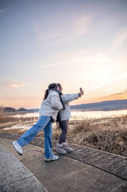 Gezinti güvertesi boyunca yürü. Deniz kenarında bir yürüyüş. Doğada yürümek. Gün batımı. Yüksek kalite fotoğraf. Sur boyunca yürüyen iki kadın. Soğuk hava. Uzayı kopyala Gün batımında fotoğraf çeken kadınlar.