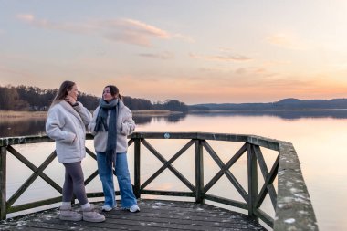 Arkadaşlar gölde gün batımını izliyorlar. Gün batımı. Doğada yürümek. İskele. Berth. Yüksek kalite fotoğraf. İskelede gün batımını izleyen iki kadın. Rıhtımda iki kadın. Boşluğu kopyala.