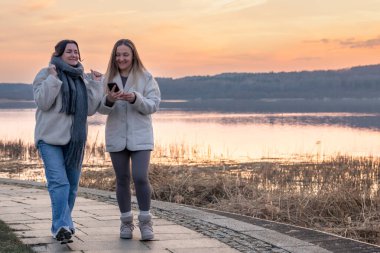 Gezinti güvertesi boyunca yürü. Deniz kenarında bir yürüyüş. Doğada yürümek. Gün batımı. Yüksek kalite fotoğraf. Sur boyunca yürüyen iki kadın. Soğuk hava. Boşluğu kopyala.