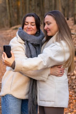 Ormanda telefonlarıyla selfie çeken iki arkadaş. Doğadaki arkadaşlar. Yüksek kalite fotoğraf. İlkbahar, sonbahar mevsimi. Ormanda bir yürüyüş, doğada..