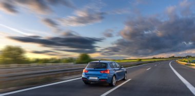 Kırmızı bir araba gün batımında doğada bir otoyolda ilerliyor. Arka planda rüzgar türbinleri ve dinamik bir gökyüzü var..