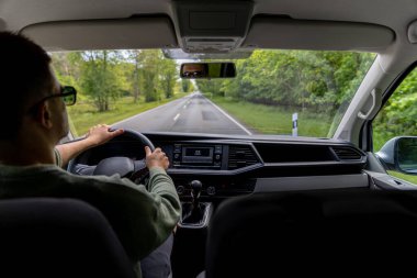Bir adam, ormanla çevrili kırsal yay yolunda dikkatle araba sürer. Yolcu tarafından çekilen görüntü, sakin hareket ve sessiz gözlemi yansıtıyor..