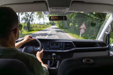 Güneşli bir günde yeşil ağaçlarla çevrili kırsal bir asfalt yolda bir sürücü, arabanın içinden yakalanır..