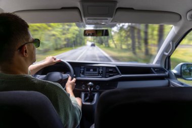 Güneş gözlüklü bir adam bir yaz günü düz bir otoyol boyunca araba sürer. Dışarıdaki yeşil manzara sakin yol atmosferini arttırıyor..