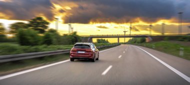 Kırmızı araba gün batımında otoyolda hızla ilerliyor. Arka planda hareket bulanıklığı ve dramatik bir gökyüzü var..
