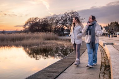 Gezinti güvertesi boyunca yürü. Deniz kenarında bir yürüyüş. Doğada yürümek. Gün batımı. Yüksek kalite fotoğraf. Sur boyunca yürüyen iki kadın. Soğuk hava. Boşluğu kopyala.