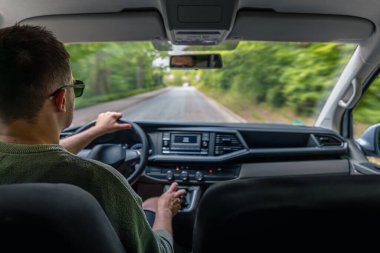 Güneş gözlüğü takan bir adam kırsal bir yolda araba kullanıyor. Pencerenin önünden geçen ağaçlarla iç mekan manzaralı..