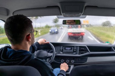 Yağmurlu bir yolda giden bir arabanın iç manzarası, direksiyonun arkasındaki sürücü, önünde yağmur damlaları ve arabalar olan ıslak ön cam..