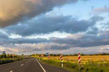 Asfaltlı bir yol, akşam güneşiyle aydınlanan bulutlarla dolu dinamik bir gökyüzünün altında, açık bir arazi boyunca uzanır..