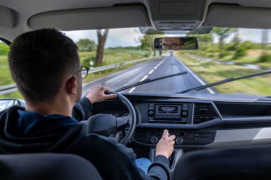 Ön koltukta kapüşonlu bir kız ve araba kullanan bir adam var. Yağmurlu yol ve yeşil ağaçlar ön camdan görülebilir..