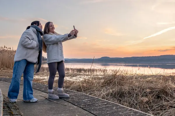 Gezinti güvertesi boyunca yürü. Deniz kenarında bir yürüyüş. Doğada yürümek. Gün batımı. Yüksek kalite fotoğraf. Sur boyunca yürüyen iki kadın. Soğuk hava. Uzayı kopyala Gün batımında fotoğraf çeken kadınlar.