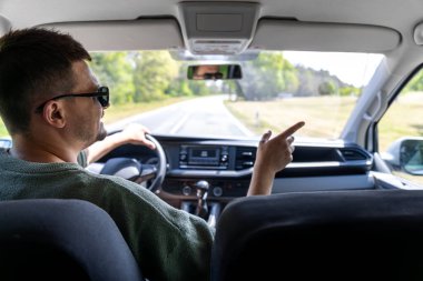 Güneş gözlüklü bir adam orman yolunda araba sürüyor. Geniş kabin manzarası hem sürücüyü hem de manzaralı kırsal bölgeyi ön camdan yakalar..