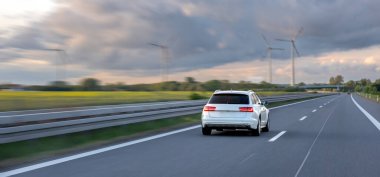 Bulutlu bir akşamda arka planda rüzgar türbinleriyle otoyolda giden beyaz bir araba..