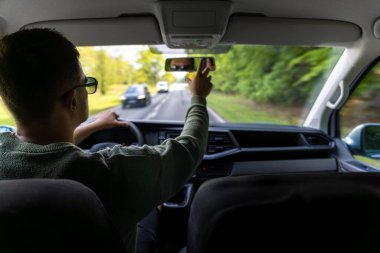 Güneş gözlüklü bir adam bahar aylarında dolambaçlı bir orman yolunda araba sürer. Ön koltuk görüntüsü yolculukta dalmayı ve sürekli kontrolü yakalar..