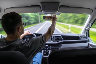 Adam yeşil orman yolunda araba sürerken dikiz aynasını ayarlar..