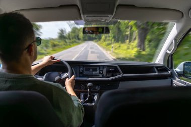 Güneş gözlüklü bir adam, yeşil bir yaz günü çevresi yeşilliklerle çevrili düz bir kırsal yolda araç kullanıyor..