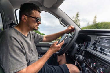 Güneş gözlüklü bir adam araba sürerken, bir eli direksiyonda, gerçekçi bir iç mekanda akıllı telefona yoğun bir şekilde bakar..