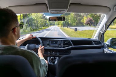 Güneş gözlüklü bir adam parlak bir yaz gününde otoyol boyunca araba sürer. Pencerenin dışındaki doğa ile iç görünüm.