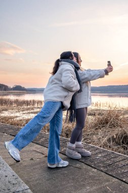 Gezinti güvertesi boyunca yürü. Deniz kenarında bir yürüyüş. Doğada yürümek. Gün batımı. Yüksek kalite fotoğraf. Sur boyunca yürüyen iki kadın. Soğuk hava. Gün batımında fotoğraf çeken kadınlar.