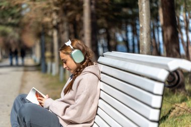 Parkta kitap okuyan genç bir kız bankta oturuyor. Parkta kitap okuyan bir kız. Yüksek kalite fotoğraf. Mesaj için yer var. Kulaklıklı kız kitap okuyor..