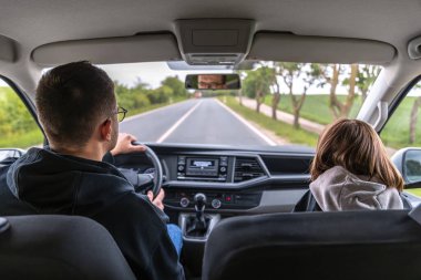 Bir adam yeşil orman yolundan geçiyor, ön camdan görülüyor. İçerisi sakin ve odaklanmış gibi, yolda son zamanlarda yağan yağmurun izleriyle..