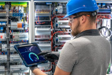 Bir kask ve eldiven uzmanı karmaşık bir santralin önünde dururken tablet ekranı işaret ediyor. Endüstriyel bir iç mekanda dijital tanı sahnesi.