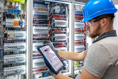 Koruyucu giysili bir adam, santralin yanındaki tablette bulunan elektrik şemasını analiz ediyor. Dijital araçlar, modern teknik ortam, yapay ışıklandırma.