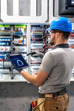 Koruyucu giysili bir adam elektrik panelinin önündeki tabletten veri okuyor. Bir santral odasında dijital sistem denetimi sahnesi.