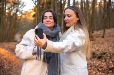 Ormanda telefonlarıyla selfie çeken iki arkadaş. Doğadaki arkadaşlar. Yüksek kalite fotoğraf. İlkbahar, sonbahar mevsimi. Ormanda bir yürüyüş, doğada..
