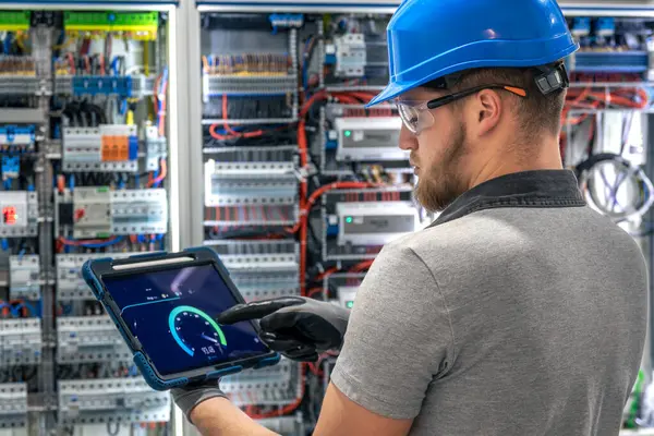 Bir kask ve eldiven uzmanı karmaşık bir santralin önünde dururken tablet ekranı işaret ediyor. Endüstriyel bir iç mekanda dijital tanı sahnesi.