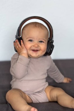 Geniş tebessümlü bir bebek kulaklıklarını kulaklıklarına dayar, rahat bir iç mekanda bir yatakta oturur..