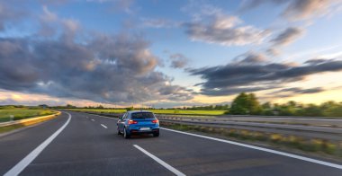 Kırmızı bir araba, bulutlu bir günbatımı gökyüzünün altında yol boyunca hızla ilerliyor. Özgürlük ve hareket çağrıştırıyor..