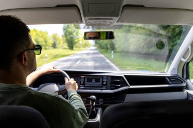 Bir adam bahar manzarasında araba sürerken dikiz aynasını ayarlar. Çerçeve elini, aynayı, kabinin bir kısmını ve gün ışığını yakalar..