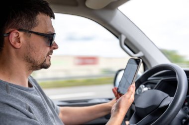 Adam ekranda navigasyon uygulaması olan akıllı telefonuna bakarken araba kullanıyor. Parlak gün ışığı ve hareket pencerelerden görülebilir..