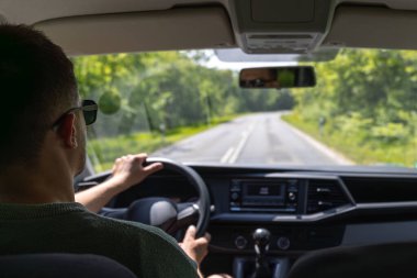 Koyu renk güneş gözlüğü takan bir adam, güneşli asfalt bir yolda orman yeşilliği ve iç perspektifle kaplı bir şekilde araba sürer..