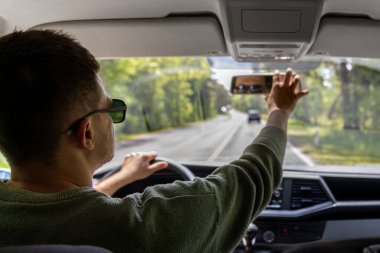 Güneş gözlüklü bir adam kırsal bir yolda araba sürerken dikiz aynasını ayarlar. Kabin manzarası bir bahar gezisi sırasında odak ve güvenliği vurgular.