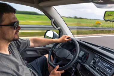 Güneş gözlüklü bir adam kırsal bir otoyolda güvenli bir şekilde araba sürer. Hareket bulanıklığı seyahat sahnesine dinamik ekler.