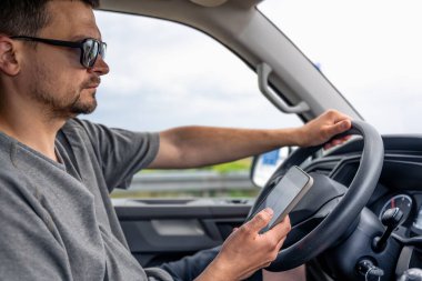 Güneş gözlüklü adam araba sürerken bir akıllı telefon tutuyor. Günlük araba kullanımından gerçekçi ve modern bir sahne.