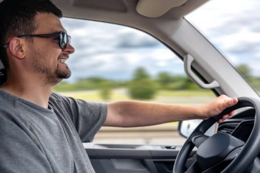 Güneş gözlüklü bir adam yüzünde bir gülümsemeyle araba sürer. Bulanık arkaplan hareket ve rahatlamış seyahat hissini arttırır..