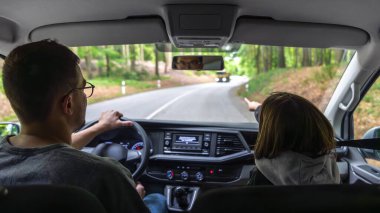 Çocuk, yetişkin bir sürücüyle orman yolunda giderken ön koltuktan el kol hareketi yapar..