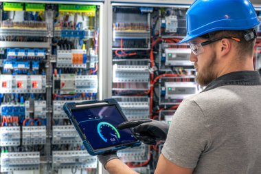 Güvenlik teçhizatı kullanan bir teknisyen, şebeke hızını tablet ile santralin önünde belirler. Endüstriyel bir iç mekanda dijital kontrol.