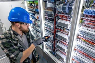 Koruyucu giysili bir adam elinde tornavidayla elektrik panelinin önünde duruyor..