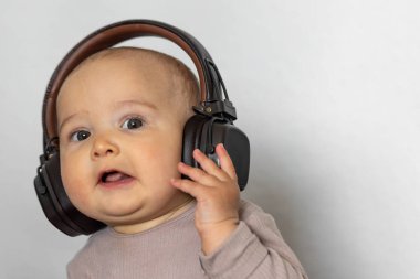 Aşırı büyük kulaklık takan ve kameraya tuhaf bir şekilde bakan bir bebeğin yakın plan portresi. Yumuşak ışıklandırma ve nötr bir arkaplan duygusal hissi arttırır..