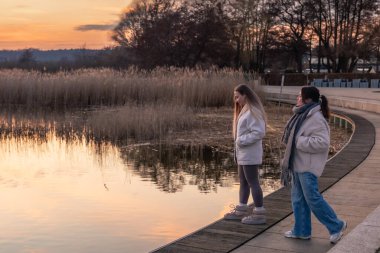 Gezinti güvertesi boyunca yürü. Deniz kenarında bir yürüyüş. Doğada yürümek. Gün batımı. Yüksek kalite fotoğraf. Sur boyunca yürüyen iki kadın. Soğuk hava. Boşluğu kopyala.