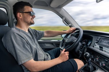 Güneş gözlüklü bir adam, iki eli direksiyonda, gülümseyen modern bir araba kullanır. Çekim iç detayları ve rahat bir sürüş anını vurguluyor..